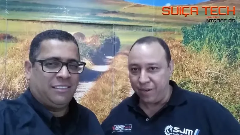 Dois homens de camisa azul-marinho sorriem para a câmera, diante de um painel com a foto de uma paisagem de campo.