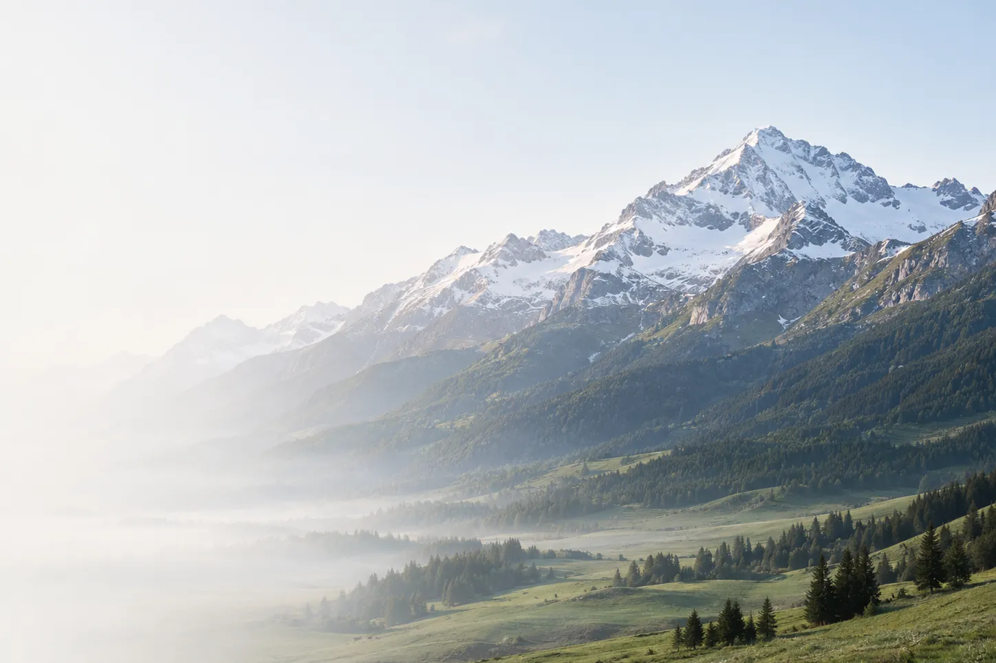 Um vale dos Alpes numa manhã clara, com picos nevados ao fundo e névoa sobre os campos.